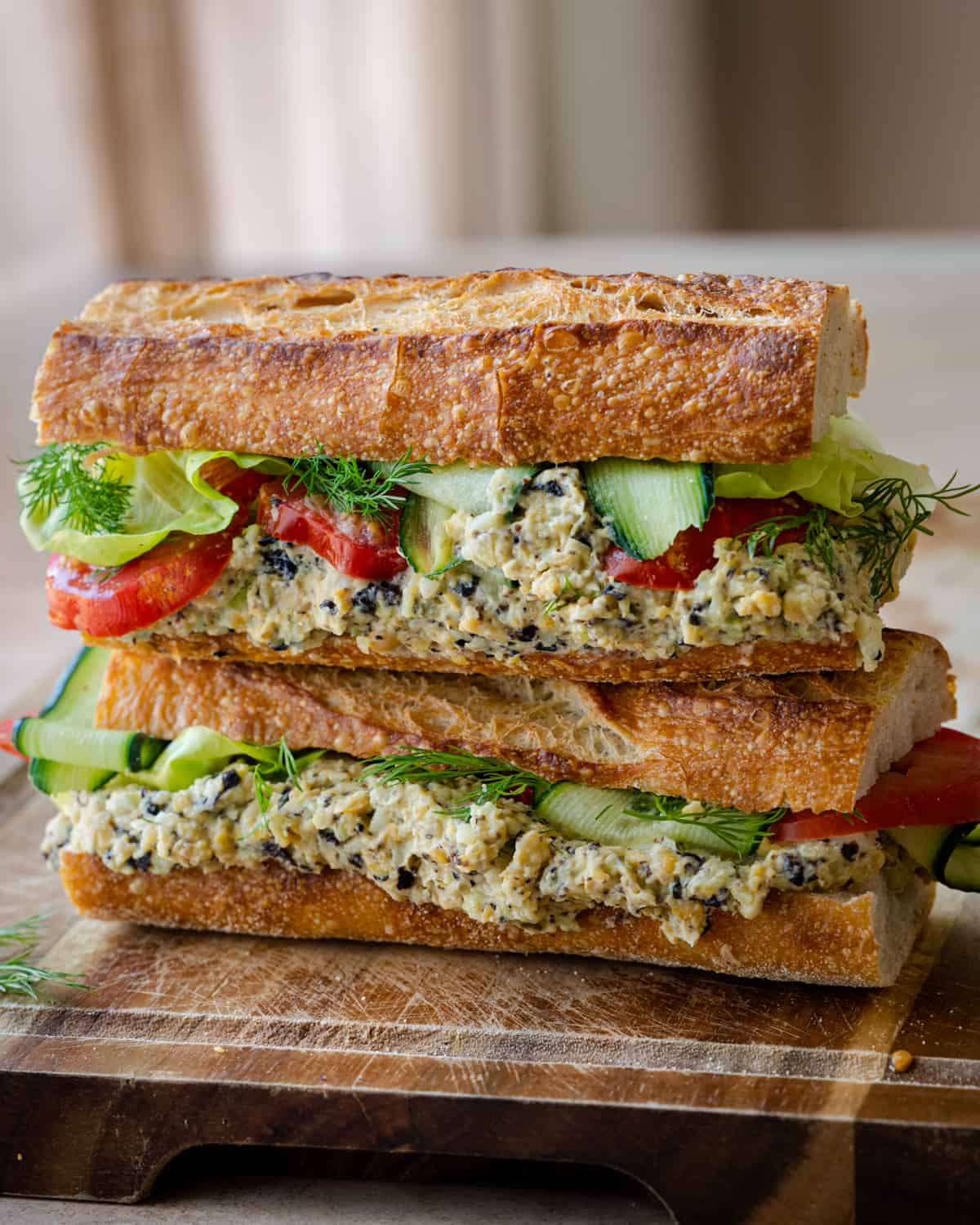 two baguette sandwiches stuffed with vegan tuna salad, cucumber, lettuce and tomato on wooden board.