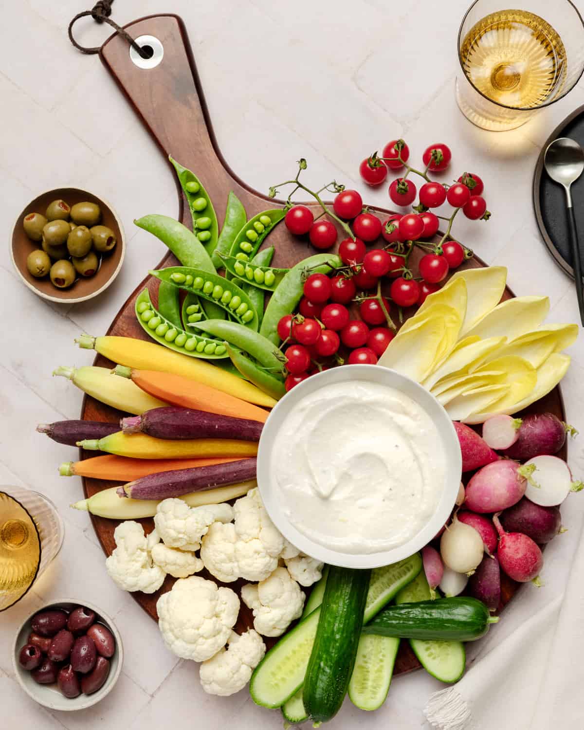 Aioli with various vegetables on a round wooden serving board on a white table.