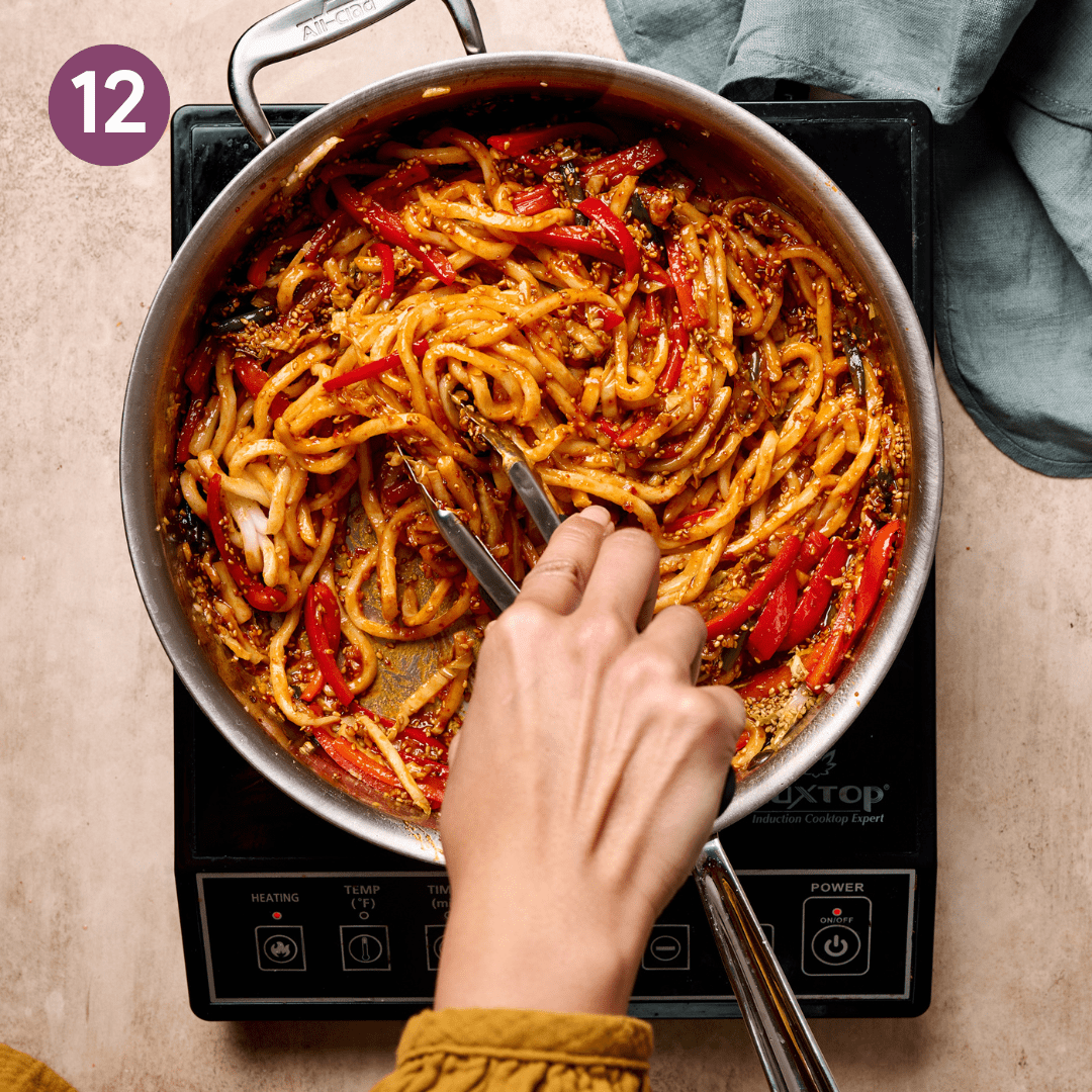 Person using tongs to stir udon noodles into saucy veggie mixture.