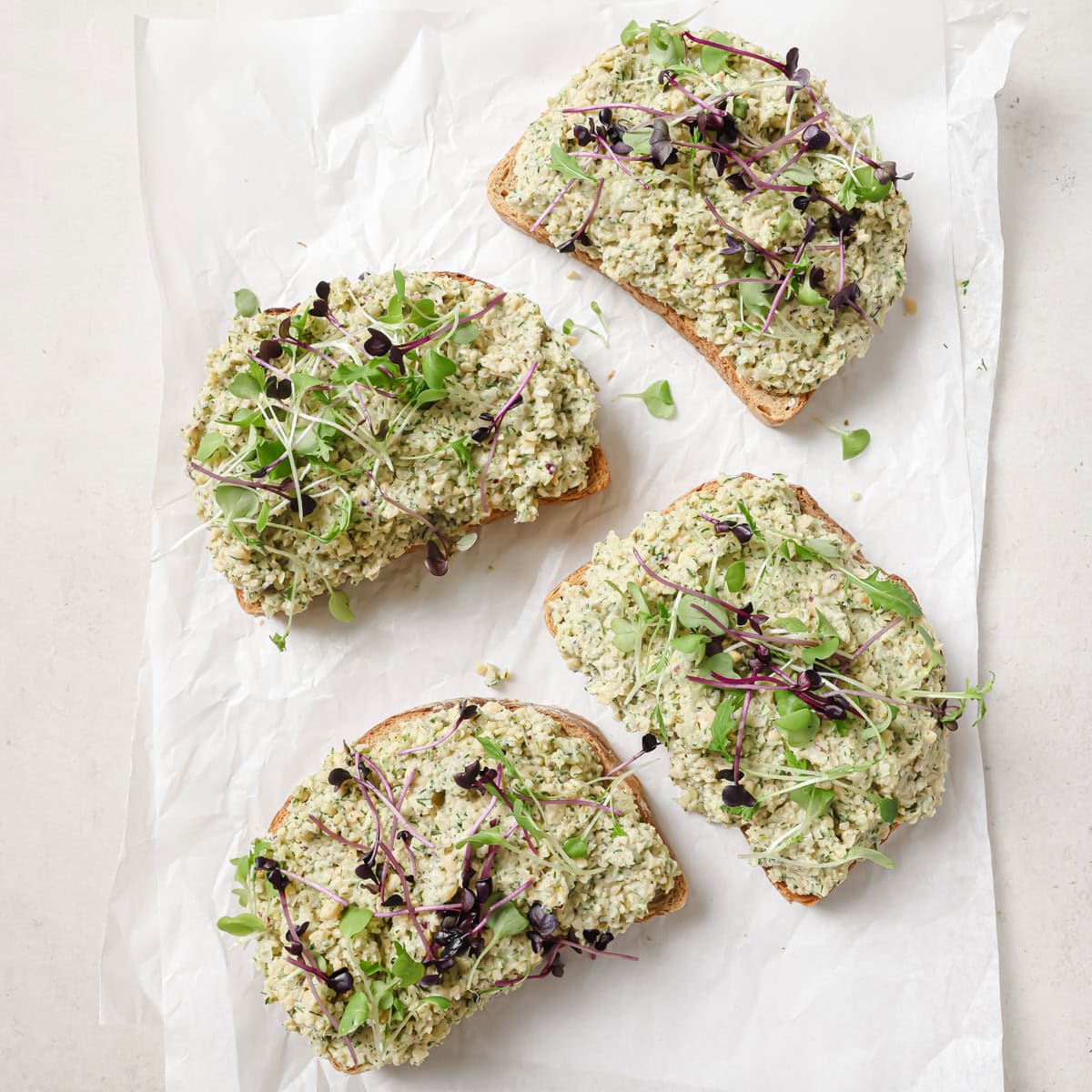 four pieces of toasted bread with chickpea salad and microgreens on a piece of parchment paper.