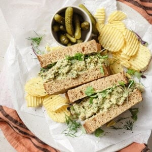 two chickpea salad sandwich halves on a plate with potato chips and cornichons.
