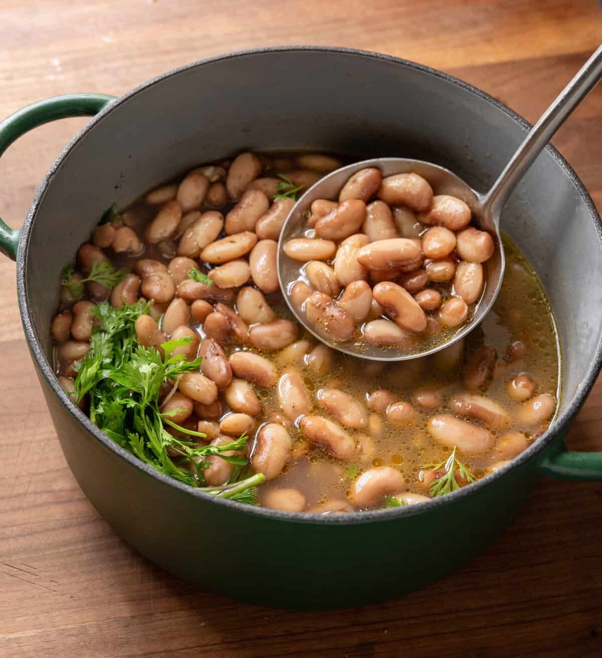 ladling brothy beans into a green saucepan on a wooden table.